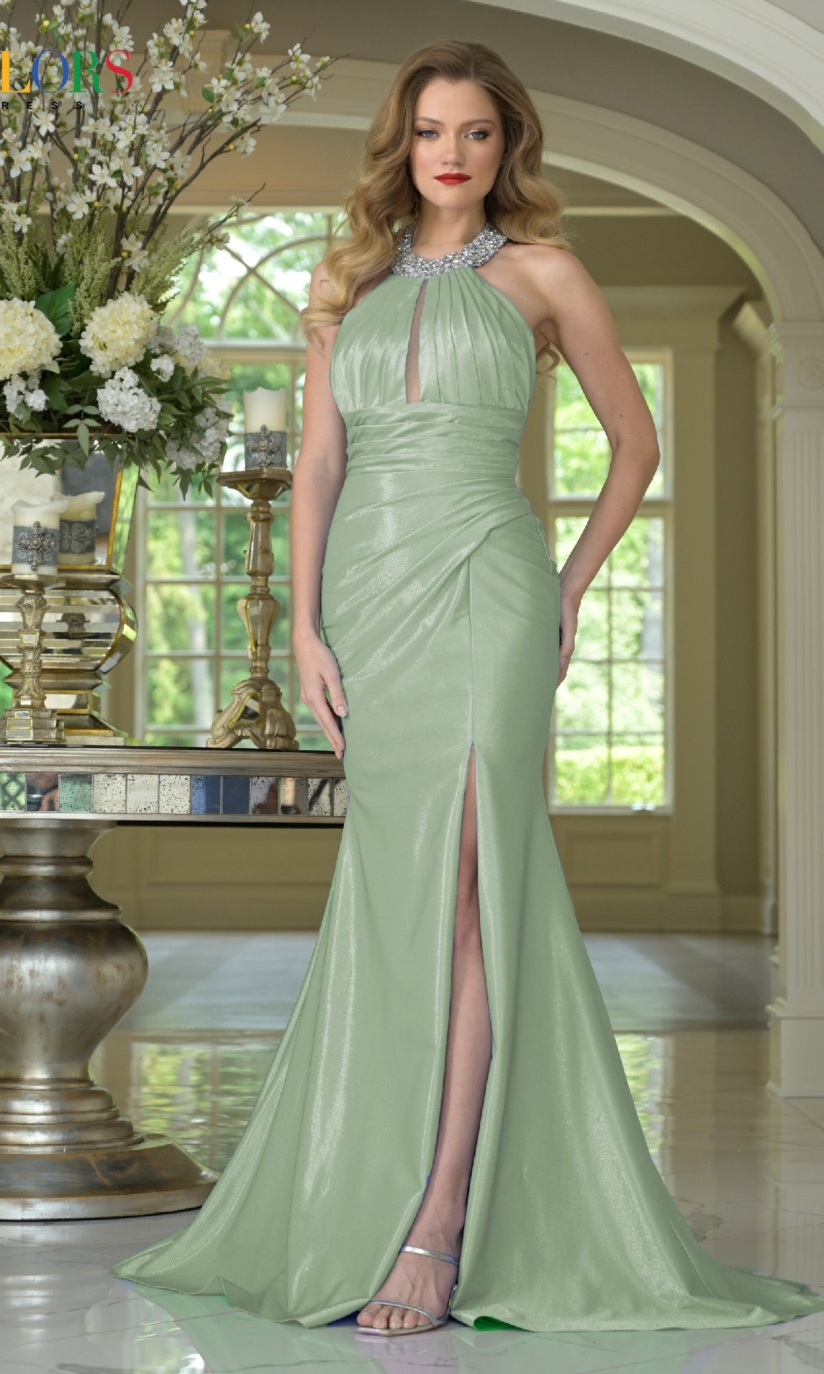 A woman poses indoors beside floral arrangements wearing the Colors Dress 3835, a light green halter fitted long prom dress with an embellished high neckline, keyhole cutout, thigh-high front slit, and styled hair.